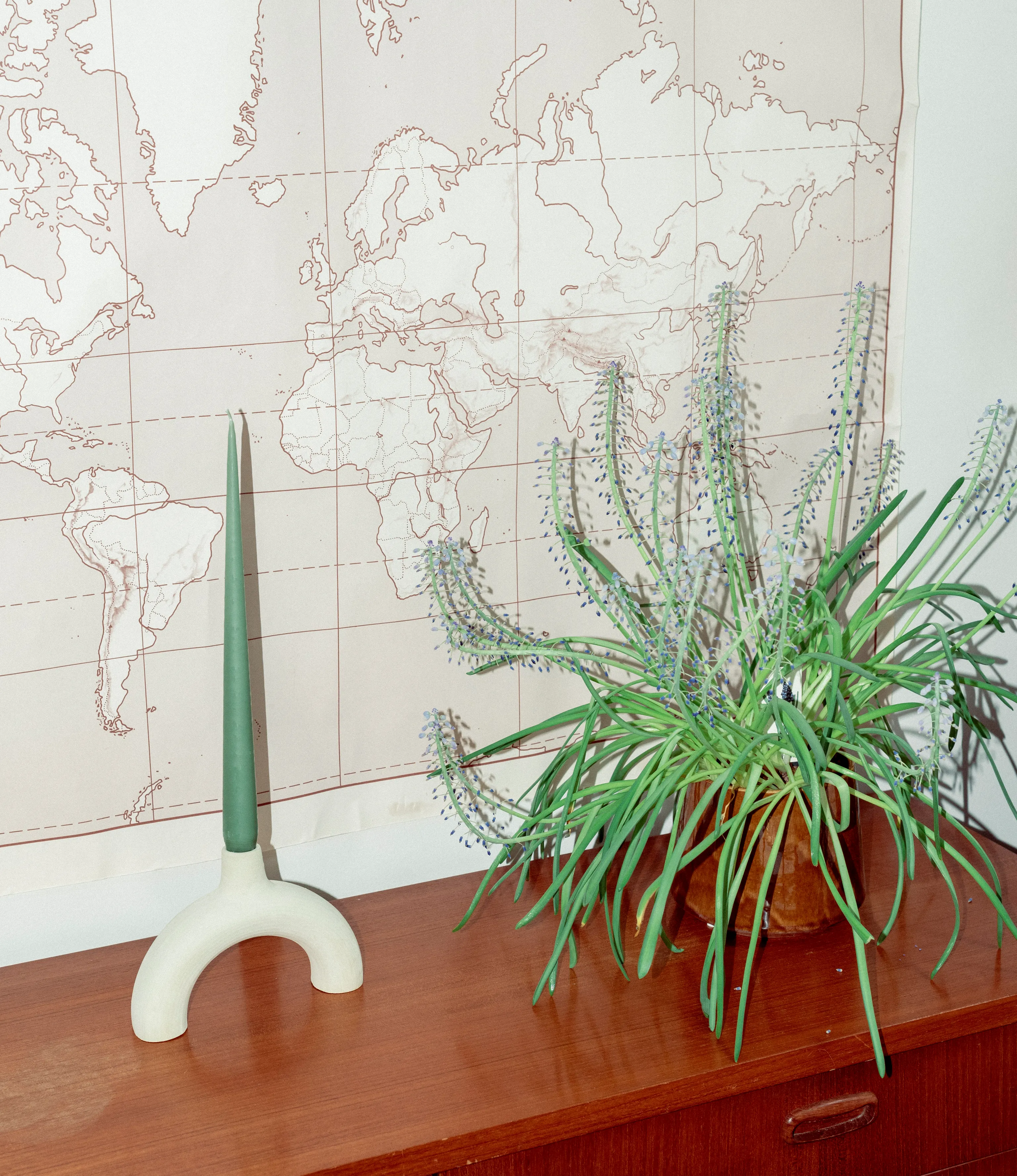 Arc Candle Holder from Alfareria La Nava comes in the color raw white. It has a matt, raw finishing. In this picture you can see the product placed on a drawer next to a plant and a map. The product is in use with taper candle coming in the soft eucaliptus color hue.