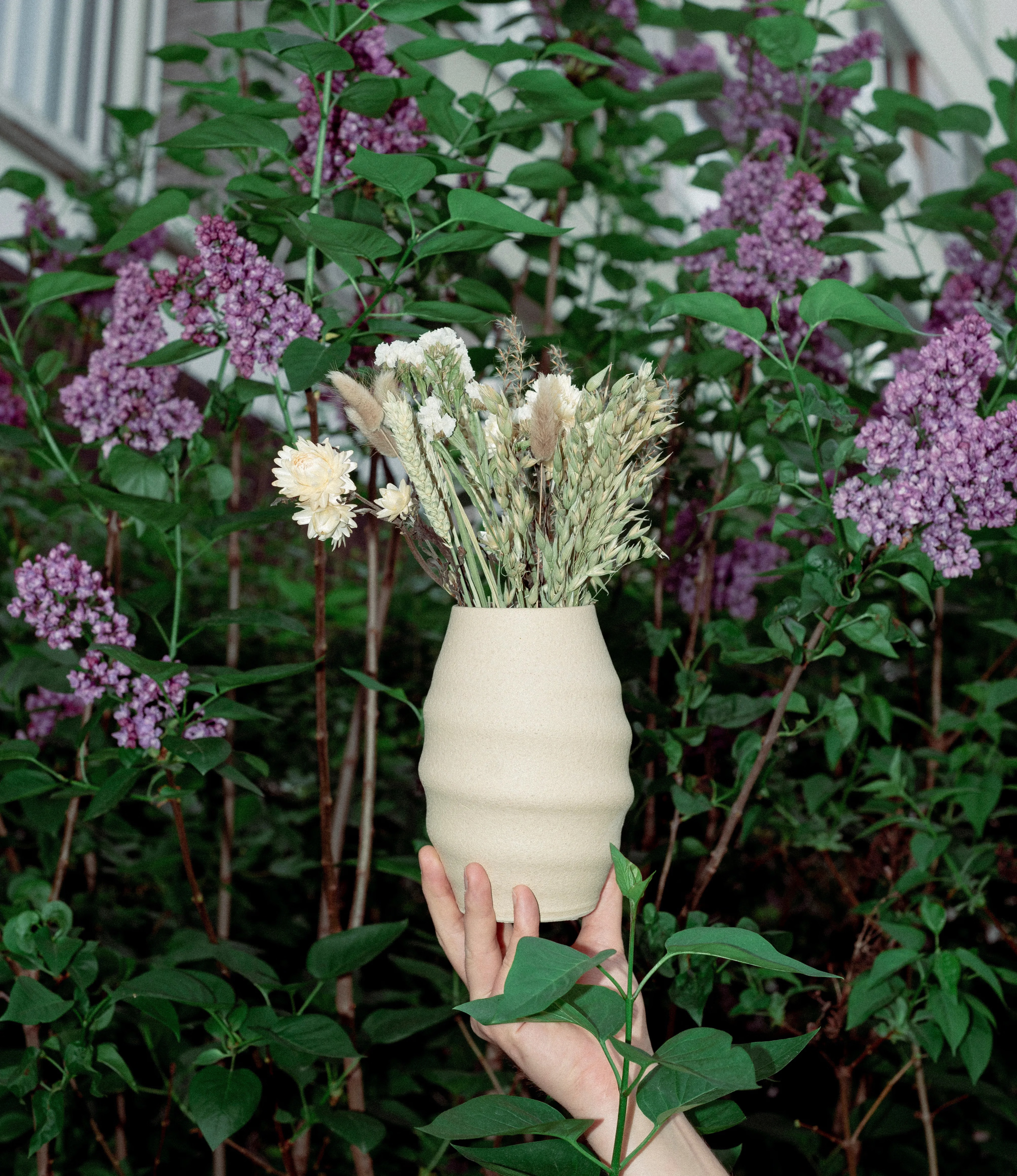 This is a picture of Aonia Vase in the nature hold by a hand. In the background there are lilac flowers and in the vase there is a dried flower bouquet.