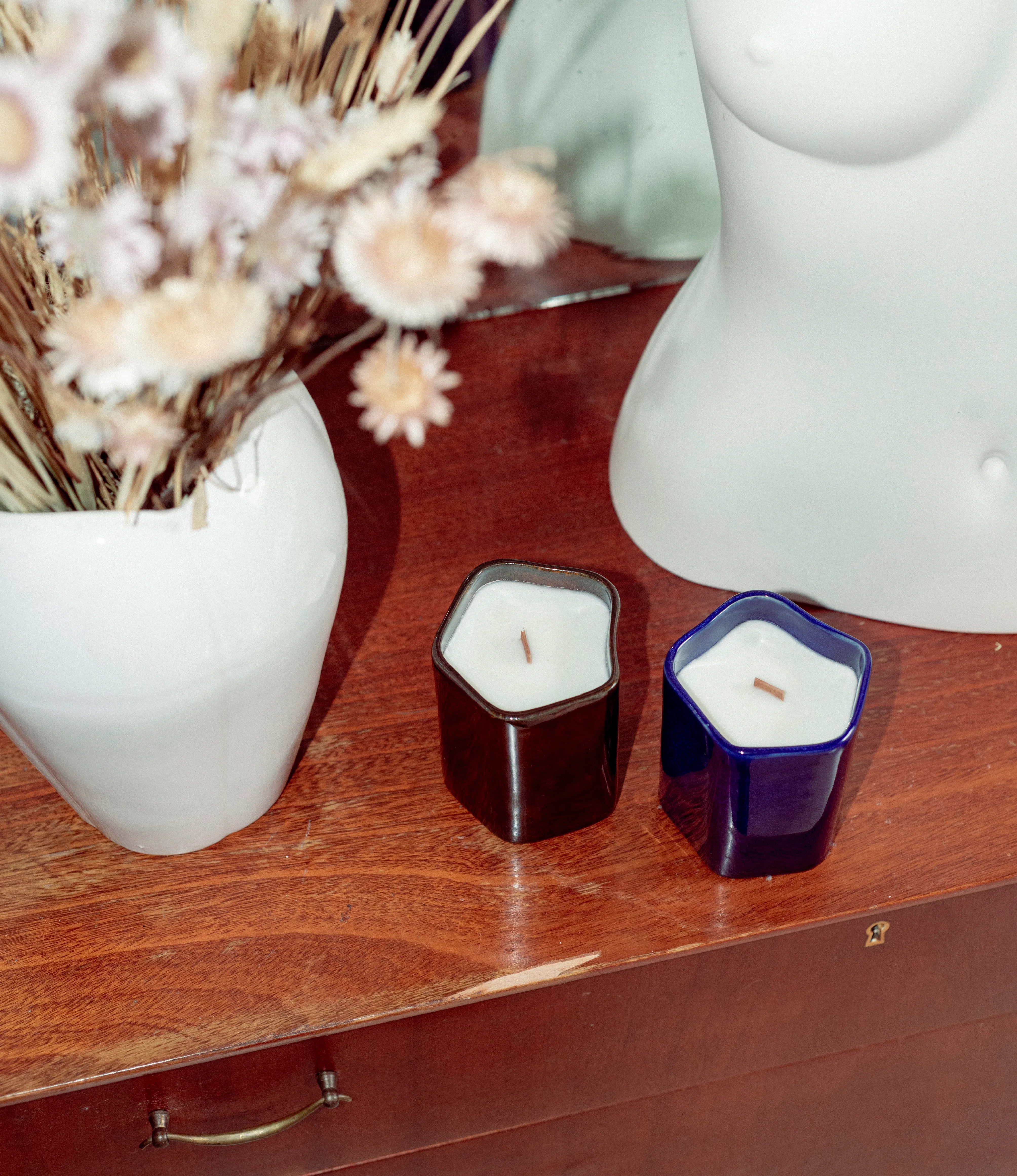 Two of the Ocactuu's scented wax candles are showcased on top of a drawer, next to a bouquet of dried flowers.
